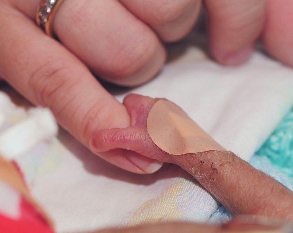 Mała dłoń wcześniaka z plasterkiem, zaciskająca się na palcu dorosłego – symbol walki o życie, którą wspiera Twoje 1,5% podatku.