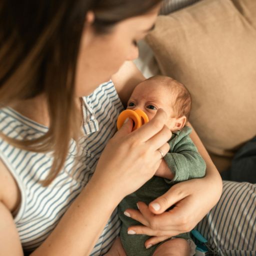 Kobieta w bluzce w poziome paski trzyma noworodka w zielonym body i błękitnej czapeczce, delikatnie wkładając mu do ust mały, pomarańczowy smoczek. W rozmytości tła widać szpitalne wyposażenie, w tym inkubator. Miękkie, naturalne światło oświetla scenę.
